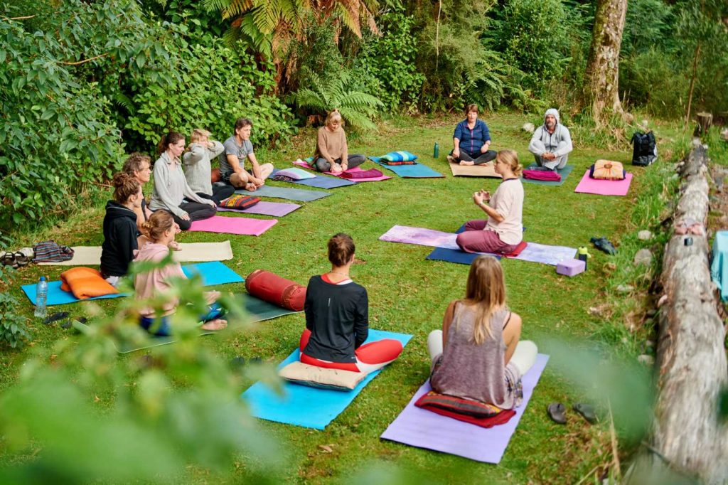 A group of humans sitting cross-legged on yoga mats laid in a semi circle on a grassy lawn with Kate leading from the centre. They are meditating before beginning a yoga sequence.